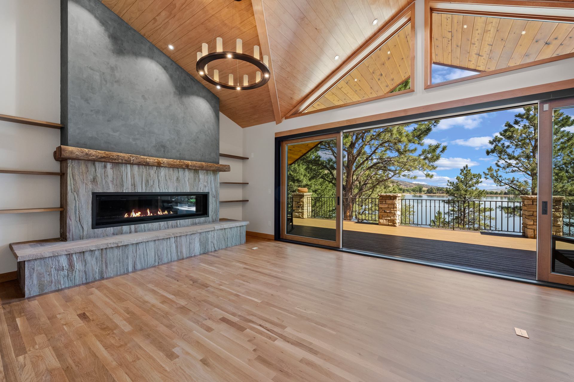 Living room with linear gas fireplace, stone surround, reclaimed-timber mantel, and glass wall to the lake deck