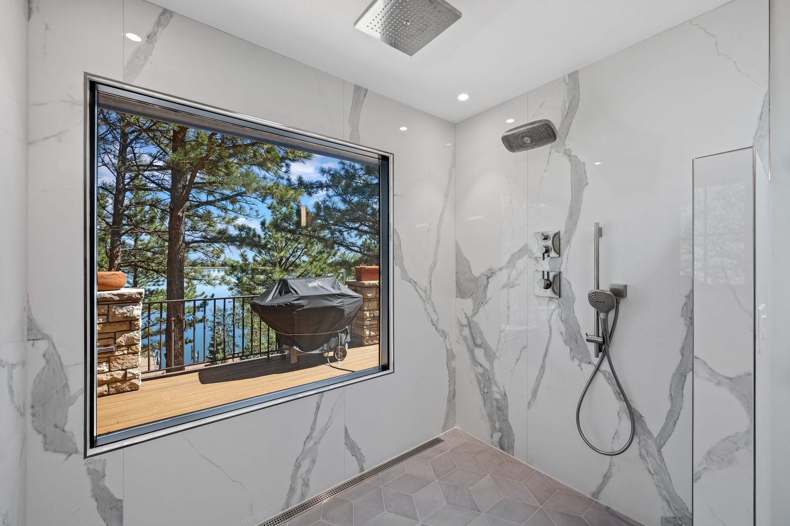 Marble walk-in shower with rain head, handheld sprayer, and a picture window framing pine trees and water